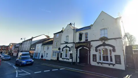 Google A Google Maps streetview picture of the Steam Railway pub. The building is double fronted with two pointed roofs, rendered in cream plaster with a black sign over the front door which reads 'Steam Railways Co' in gold lettering. 