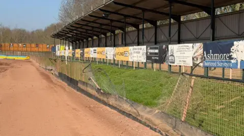 Leicester Lions Speedway Broken fencing with stadium behind and sandy track in front.