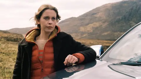 Black Camel Pictures/BBC Alba/John Maher Star Sorcha Groundsell as Kat in An t-Eilean (The Island) - she is outside leaning on a car bonnet, with the hills pictured behind her. She has auburn hair, a pensive look on her face and a brown and orange jacket on.