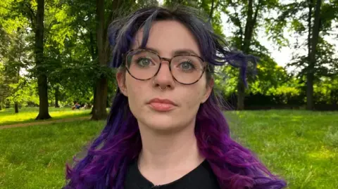 A woman with shoulder length purple hair, and tortoiseshell glasses, and two nose rings, looks at the camera. She's standing in a park with trees and grass behind her, and is wearing a black top. 