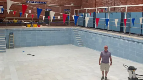 Brian McCann wearing a purple and white striped T-shirt and cream shorts stands smiling in the middle of the empty pool Pacific Pool with a smile on his face. Hanging above him and the pool are multi-coloured backstroke flags. There are tools and equiptment on the side of the pool. 