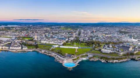 An aerial shot of Plymouth city centre showing the Tinside Lido and Smeaton's Tower in the foreground and views out to Dartmoor in the distance.