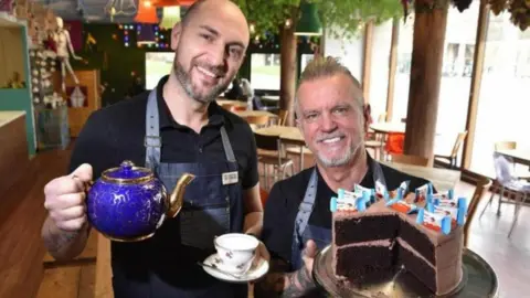 Two men in a cafe. The one on the left is holding a tea cup and saucer in one hand a bright blue and gold teapot in the other hand. The man on the right is holding a large iced chocolate cake on a plate