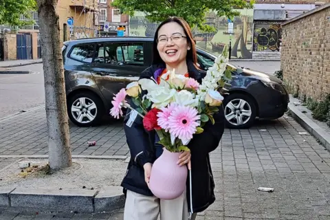Metropolitan Police Service A woman with long dark hair and wearing glasses is standing outside. She is smiling and holding a large pink vase filled with brightly coloured flowers. There is a tree trunk on the left-side of the photo, and a car in the background.