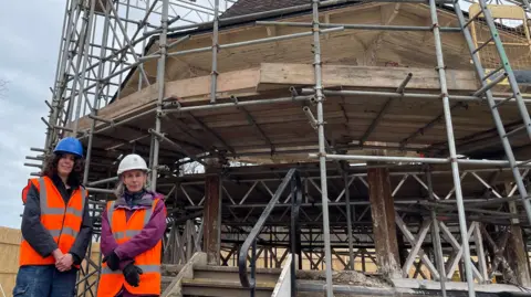 Emma Hampton on the left is looking directly at the camera and smiling. She is wearing a black jacket with an orange fluorescent hi-vis vest over the top and blue hard hat. Next to her is Sarah Morrison, a woman who is also looking directly at the camera and she is wearing a purple outdoor jacket, orange fluorescent hi-vis vest and white hard hat. Behind them is a bandstand covered in scaffolding while restoration works are carried out.