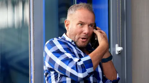 PA Derek Hatton with a checked blue shirt is on a mobile phone