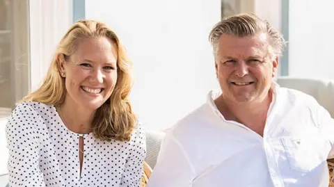 The Moorings Hotel The image shows Iselin and Matthew Jones sitting down and smiling at the camera. Iselin is wearing a white blouse with black dots on it. Matthew is wearing a white Oxford shirt with a breast pocket.