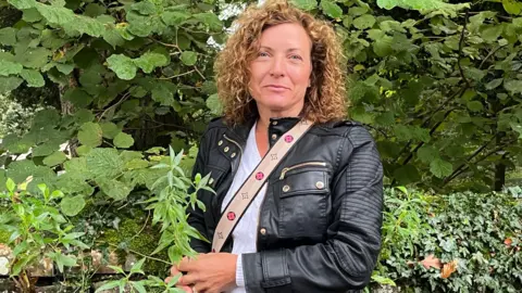 Julia Gregory/BBC Sally Pavey has shoulder length curly hair and is standing outside surrounded by greenery. She is looking at the camera and wearing a black jacket and white top with a bag strap over her shoulder.