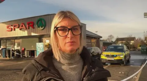 Sinn Féin assembly member Linda Dillon standing outside a police cordon at a petrol station in Clone, County Tyrone. She has blonde hair, black-rimmed glasses and a black padded coat over a grey knitted turtle neck