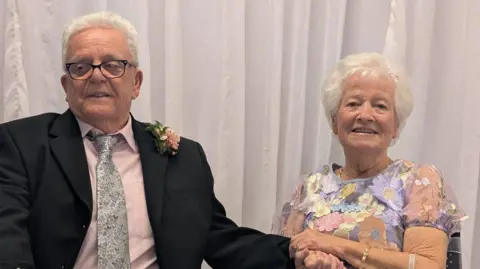Contributed Terry and Jo sit in chairs in a registry office. Terry wears a black suit, pink shirt, blue floral tie and glasses. He looks away from the camera and has one hand in Jo's. Jo wears a light purple dress with floral patterns on it. She is smiling at the camera.