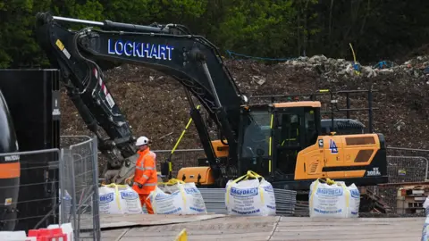 A worker in orange overalls stands next to a mound of waste which appears to tower above a nearby large mechanical digger.