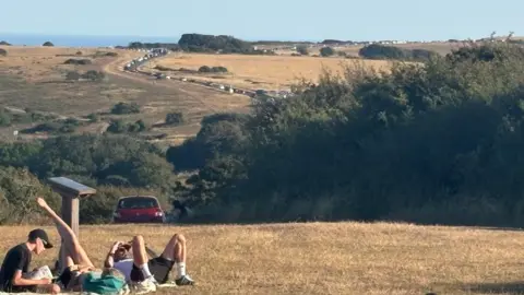 BBC / Hanna Neter Several people sit and lie on dry grass on high ground near Beachy Head in East Sussex. A long queue of cars stretches along a road beyond them, with the sea visible in the distance.