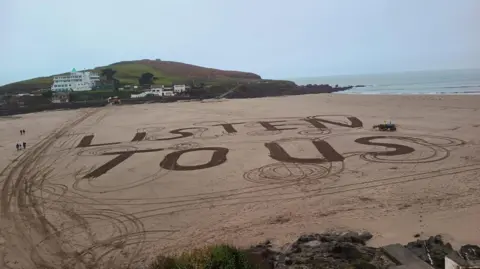 An elevated view of Bigbury Beach which has the words "Listen to us" written in giant capital letters in the sand. The beach is otherwise empty.