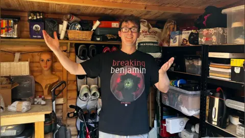 Alex Dunlop/BBC A man stands in a shed filled with goods and shelving. He is holding his arms wide open. He is wearing a T-shirt that says Breaking Benjamin.