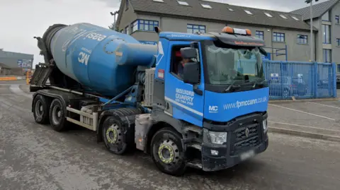 A blue concrete mixer lorry with the FP MCCann logo on it is leaving a depot. Behind the vehicle there is a large grey building.  