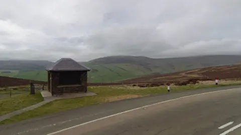 Keppel Gate, a small brown hut, is set back from the Mountain Road on the Isle of Man. The mountainous landscape, shot under cloudy skies, can be seen in the background.