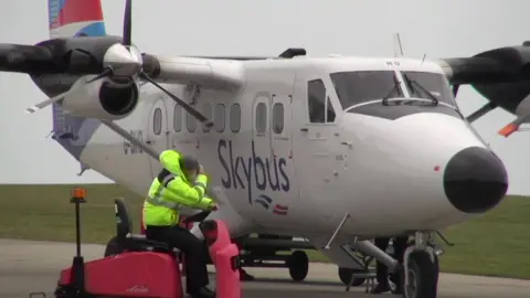 A Skybus aircraft is parked on an airport apron, with a ground crew member in a high-visibility jacket sitting on a red airport vehicle in front of it. The twin-engine turboprop plane is preparing for or undergoing routine ground operations under overcast skies.