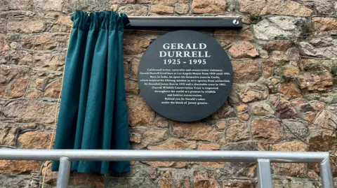 A teal curtain pulled back to unveil a new black circle plaque dedicated to Gerald Durrell on a stone wall.