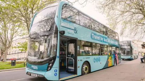 A double decker blue bus parked up on the side of a road with its door open. There are trees either side of the bus and another bus behind it. 