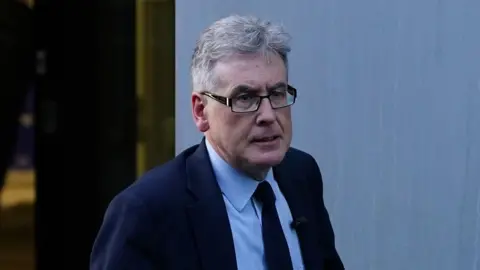 Getty Images A man with grey hair and black glasses wearing a navy suit jacket, light blue shirt and black tie