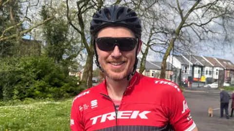 David Heathfield in Ynys Angharad Park, Pontypridd, wearing a red cycling top, helmet and sunglasses. It is a head and shoulders shot of him, and he smiles at the camera.