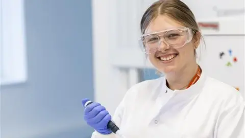 Millie-Rose Doolan in a white lab coat and blue gloves. She has dark blonde hair that has been tied back and is also wearing protective glasses. 