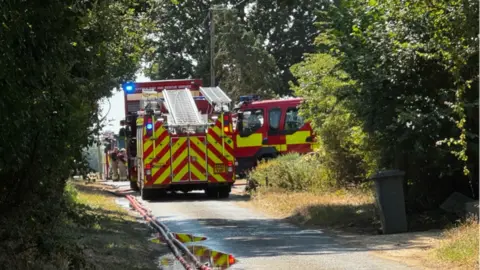 Fire engines parked along a country lane. Some firefighters are working near one of the vehicles.