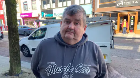 BBC Barry Mills, a Larne resident, in grey hoodie with parked traffic behind on Larne Main Street.