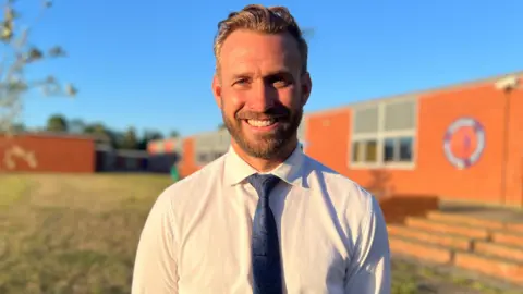 A man wearing a white shirt and navy blue tie smiling into the camera in front of a school building