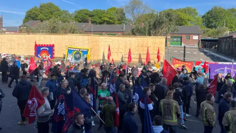 A large group of protestors gathered with large flags outside a bin depot. Some wear fire service uniforms of brown with high-vis markings. Behind them is a wooden fence with a gap in it for vehicles, which the people are blocking.