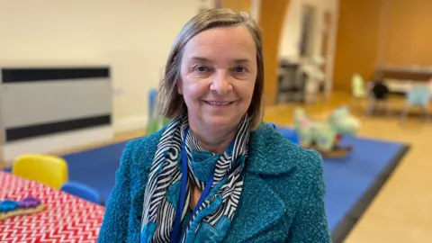 A woman wearing a blue jacket and scarf looking at the camera and smiling. There is children's play equipment and toys behind her.