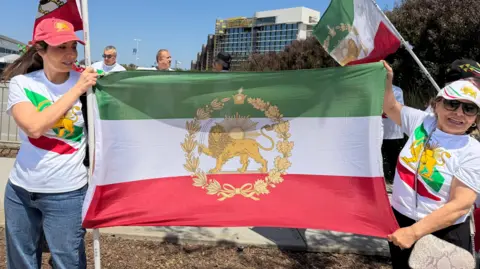 Two women wear t shirts and caps featuring the stylised lion symbol of the flag of Iran's pre-1979 Islamic revolution. They are also holding the flag between them.