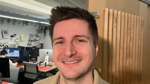 James Stafford has short dark hair and is smiling at the camera as he wears a brown coat. In the background is computer desks with someone sitting down using one.