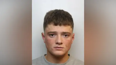 A mugshot of a young white man with brown hair and spots on his face, wearing a grey jumper.