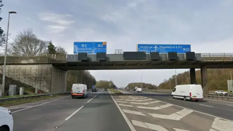 The M25 at junction 12, with blue signs over the top of the carriageway showing the way to Basingstoke, Southampton and Richmond on the M3 and towards Heathrow and Watford on the M25. The road is divided by a white chevroned section and there are some cars and vans on the road.
