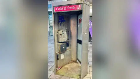 Michael Enea A phone box which is deshiveled and dirty, with a phone on the wall inside and a pink sign above which reads "coins and cards".