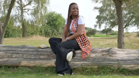 University of Wolverhampton A woman with braided hair sits on a tree trunk, which is laid out on grass. Other trees are visible behind. The woman wears a white T-shirt with a colourful jacket, black trousers and white trainers. A book with a yellow cover is placed next to her on the trunk.