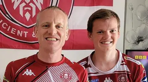 Family photograph Neil Crook smiling with his son Alex. The father and son are dressed in Wigan Warriors Rugby League kits with a Wigan Warriors flag in the background  