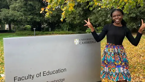 Jemimah Mensah Jemimah Mensah standing outside, in the autumn, on the right, leaning against a sign saying Faculty of Education. She has black hair pulled back behind her shoulders and is wearing a black shirt and colourful skirt. She is smiling and has raised her hands to show two V for Victory signs. 