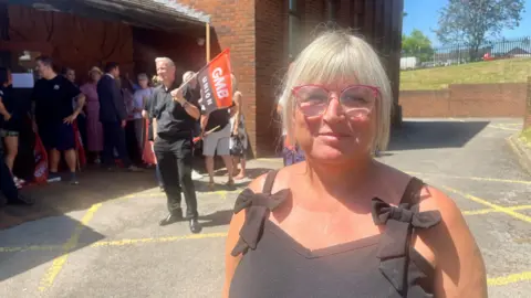 A woman with short blond hair and wearing a black top and glasses with pink frames is standing in a yard outside a brick-built building. There is a group of people gathered behind her, some carrying flags and banners.