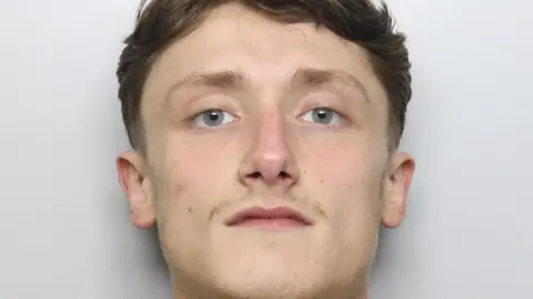 A mugshot of a 20-year-old man with blue eyes, pale skin, brown pushed back hair and a thin patchy moustache. 
