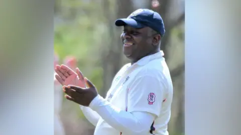 Norman Case stands on a cricket pitch, wearing cricket whites. He is also wearing a navy blue baseball cap. He is smiling and clapping his hands