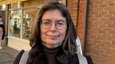 Amanda Oldershaw standing in a market place with cobbles and a bakery behind her. She is wearing a brown coat with a fur collar, has shoulder length brown hair and is wearing thinly framed glasses. She has light grey rucksack straps over her shoulders. 