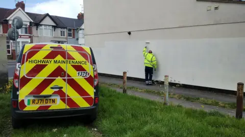 A man in a high viz jacket painting over racially motivated graffiti with white paint on the side of a house with a van and houses on a street in the background.