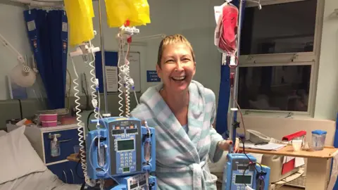 Family Paula Costin wears a light blue and white striped dressing gown and is standing in a hospital ward, flanked by various transfusion equipment. She has short light brown hair and is smiling at the camera.