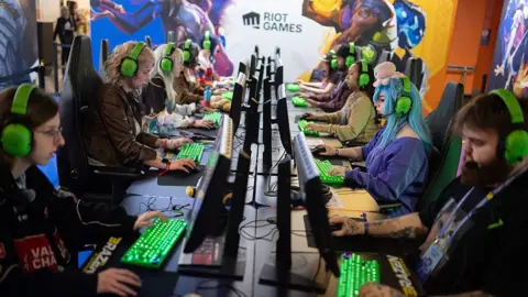 Getty Images A row of gamers sit at a long desk wearing matching green Razer headsets, focused on their screens with green-lit keyboards in front of them. A large Riot Games mural covers the wall behind them featuring colourful game characters. The players include a mix of men and women, one with blue hair and a plush toy on her head, all concentrating intently on their monitors.
