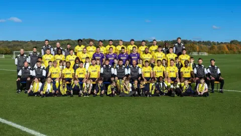 A soccer quad wearing a mix of yellow, purple and black uniforms stands in three rows on the soccer field for a photo opportunity. A row of primary school children sit on the grass at the front, wearing yellow Oxford United scarves. They say it's sunny and the sky is blue.