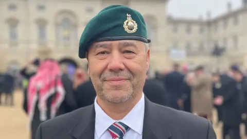 Head and shoulders of Alan Holderness smiling at the camera. He has short grey stubble and is wearing a grey suit jacker, white shirt, red and blue striped tie and green cap with a gold royal marine cap badge