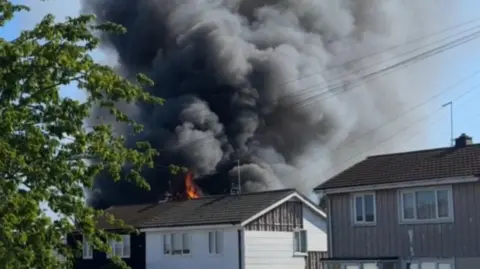 Flames and a lot of smoke behind a house in Loughton. There is a tree to the left and other houses in front. 