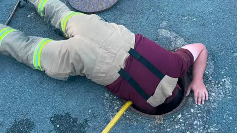 A picture of a firefighter who is leaning into a drain. His torso and legs are visible but not his head.